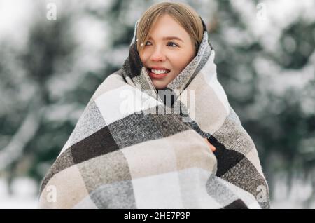 Smiling woman with warm blanket standing outdoors in winter, keeping warm. Woman covering herself with a blanket to keep warm on a snowy winter day ou Stock Photo