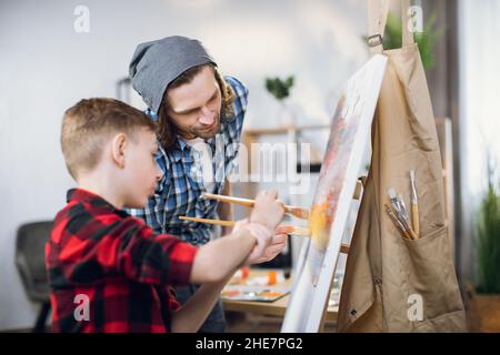 Side view of positive boy and his father using brush an colorful paints for drawing on easel. Happy family enjoying creative time spending at home. Stock Photo