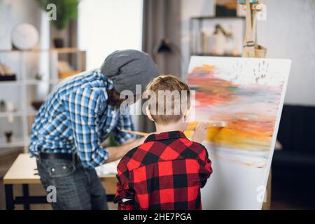 Side view of positive boy and his father using brush an colorful paints for drawing on easel. Happy family enjoying creative time spending at home. Stock Photo