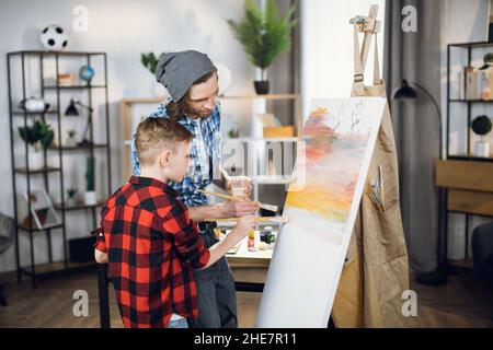 Caucasian father drawing with son with colorful paints and brush at home. Positive man and boy using white canvas for creating abstract pattern. Stock Photo