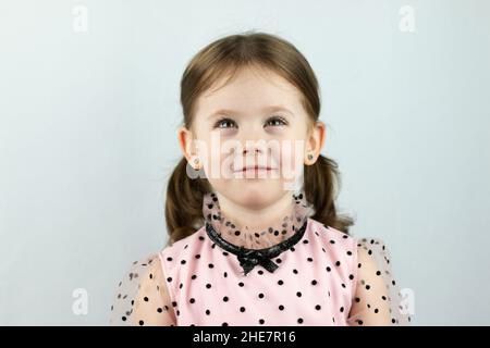 Smiling little girl with two ponytails in a dress with polka dots on a white background dreamily looks up. Studio photo Stock Photo