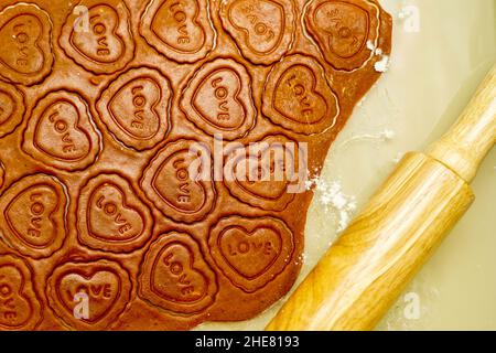 table view of making heart shaped Valentines day gingerbread cookies with the inscription love, rolling pin and rolled dough Stock Photo