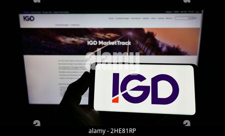 Person holding cellphone with logo of Institute of Grocery Distribution (IGD) on screen in front of business webpage. Focus on phone display. Stock Photo