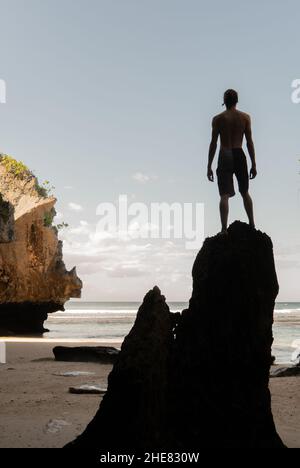 A silhouette of a young man standing on a rock on a sandy beach. Stock Photo