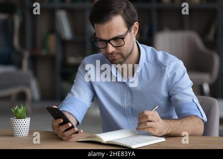 Focused confident businessman in glasses holding smartphone, taking notes Stock Photo