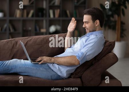 Profile view smiling man waving hand, using laptop on couch Stock Photo