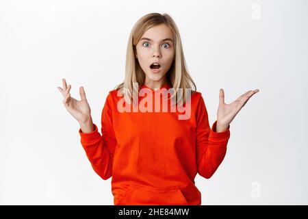 Surprised and shocked little blond girl looks down startled, seeing something lying below, drop on floor, standing over white background Stock Photo