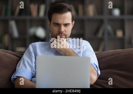 Thoughtful man looking at laptop screen, touching chin, working online Stock Photo