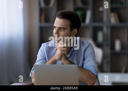 Thoughtful businessman sitting at desk with laptop, looking in distance Stock Photo