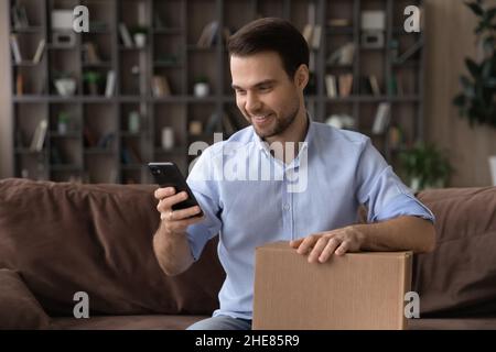 Smiling man using smartphone, holding cardboard box, unpacking parcel Stock Photo