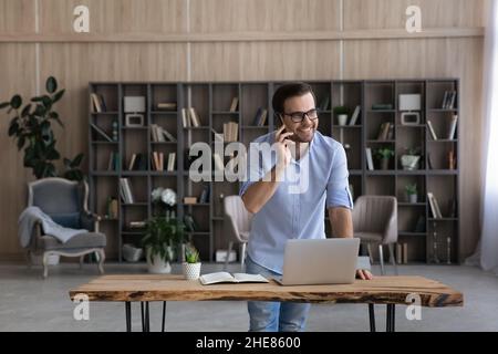 Smiling businessman in glasses talking on phone, standing near desk Stock Photo