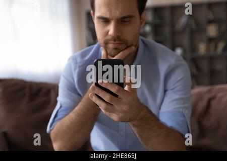 Close up thoughtful man looking at smartphone screen, touching chin Stock Photo