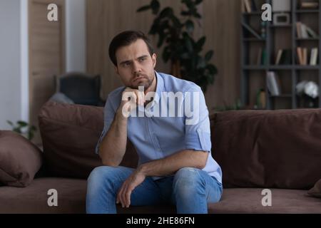 Thoughtful upset man looking in distance, sitting on couch alone Stock Photo