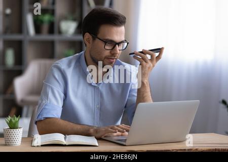 Businessman in glasses recording audio message on smartphone, using laptop Stock Photo