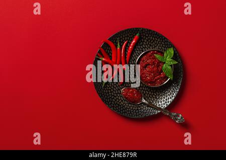 Thick hot chili sauce in a bowl and heap of fresh red hot chili peppers on a black textured plate over red background. Spices and condiments concept. Stock Photo