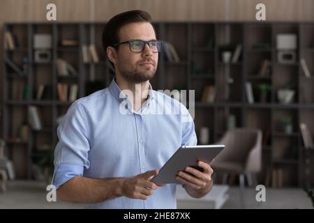 Dreamy confident man wearing glasses looking in distance, holding tablet Stock Photo