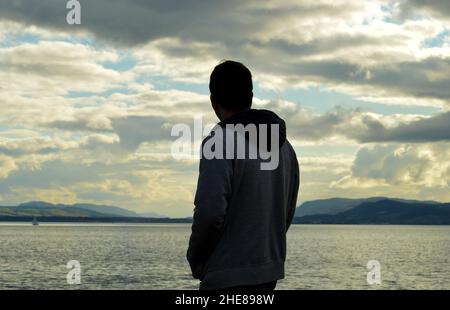 Silhouetted figure of young man looks across water to hills by Moray Firtth in Easter Ross, Scotland Stock Photo