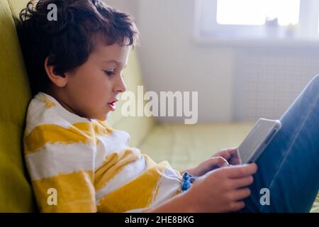 boy in yellow and white t-shirt enthusiastically plays a game on a tablet Stock Photo