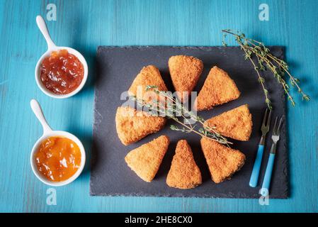 Presentation on a stone plate of fried camembert cheese next to jars with marmalade. Stock Photo