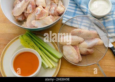 Fresh raw Chicken wings ready for deep frying with a strainer , hot sauce , blue cheese sauce and celery on a wooden cutting board in a home kitchen Stock Photo
