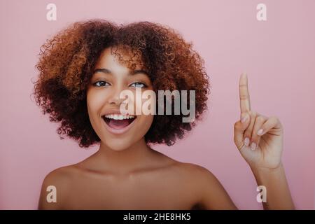 Young beautiful African-American woman with a lush curly hairstyle points her index finger up looks at the camera and smiles on a pink background, a new idea has come, inspiration Stock Photo