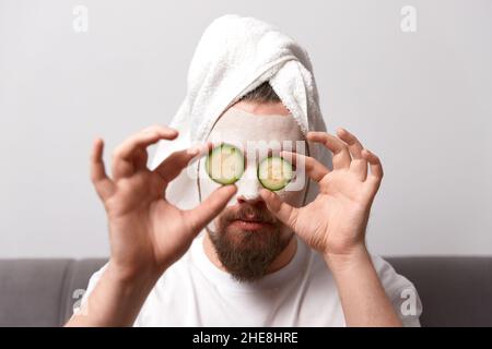 Portrait of funny happy man in white tshirt applying cucumber slices over clay mask on his face. Self care morning spa procedure Stock Photo