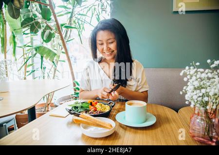 Young Asian woman with long loose black hair sits on sofa at wooden table and eats vegetable dish smiling in cosy vegan cafe closeup Stock Photo