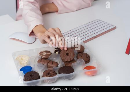England, UK. 2021. Close up of womans hand taking a chocolate biscuit from a boxed selection next to a computer keyboard. Stock Photo
