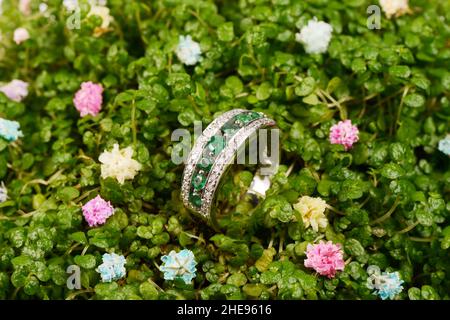 Women's platinum ring with a diamonds and blue precious emerald jemstone on micro green leaves background and polished wood, close-up. Luxury female j Stock Photo