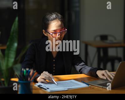 Professional female accountant with glasses working on a financial spreadsheet and laptop computer at the office. Stock Photo