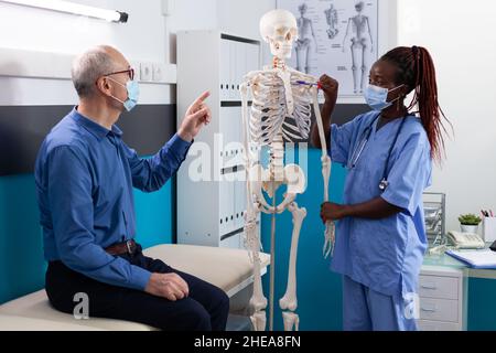 African american woman nurse with medical protective face mask against covid19 holding human skeleton model explaining spine pain to senior man patient in hospital office. Osteoporosis concept Stock Photo