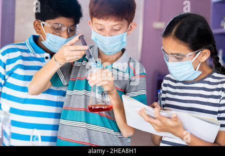 Diverse Kids experimenting with safety eyeglasses and face mask by noting results while mixing chemical using flask at chemistery laboratory - concept Stock Photo