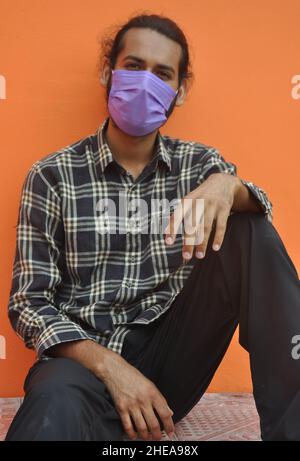 Front view of a attractive young man wearing protective face mask while sitting outside, posing against orange wall with looking at camera Stock Photo