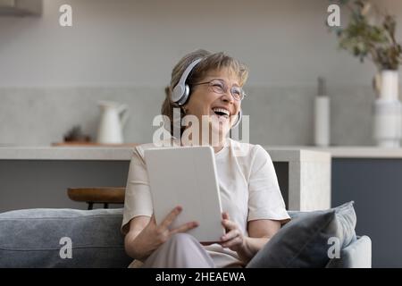 Happy elderly woman in headphones using digital touchpad gadget. Stock Photo