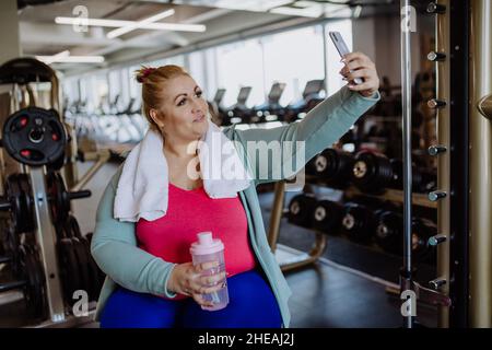 Happy mid adult overweight woman taking selfie after exercise indoors in gym Stock Photo