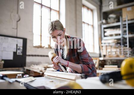 Happy female carpenter working on her product indoors in carpentry workshop. Stock Photo