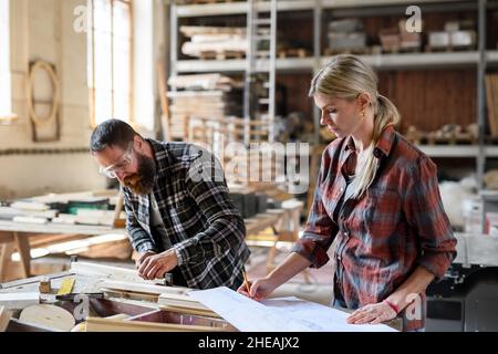 Two carpenters man and woman looking at blueprints indoors in carpentery workshop. Stock Photo