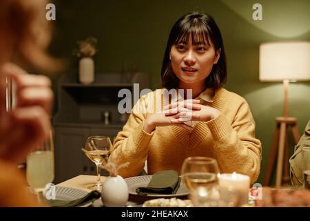 Asian young woman sitting at dining table and talking to her friends at dinner party Stock Photo