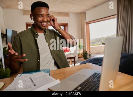 Young adult black male adjusting earphones while talking in online meeting. Sitting at kitchen counter using laptop. Stock Photo