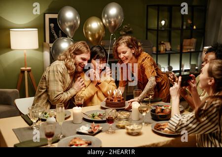 Happy young woman celebrating birthday with her friends at home, they sitting at dining table with birthday cake Stock Photo