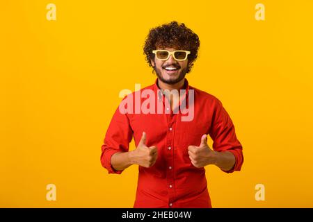 Like, excellent job. Pleased man with beard wearing sunglasses smiling to camera and gesturing thumbs up, approval hand sign, satisfied with result. Indoor studio shot isolated on orange background Stock Photo