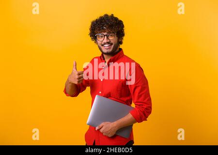 Positive smiling man freelancer with beard showing thumbs up while standing and holding laptop, satisfied with teleworking, likes his job. Indoor studio shot isolated on yellow background Stock Photo