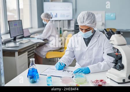 Young man in protective mask and gloves sitting at his workplace and working with diagrams at the laboratory Stock Photo