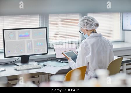 Rear view of young woman in white lab coat sitting at her workplace in front of laptop and using digital tablet at her work Stock Photo