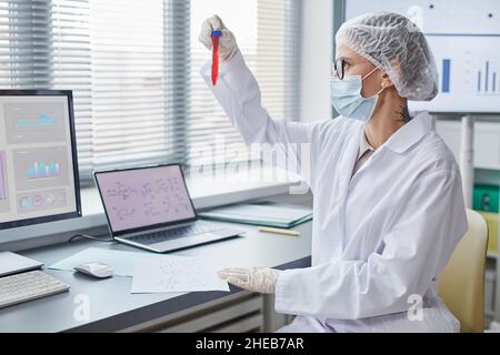 Female medical researcher examining analysis in test tube sitting at the table in front of computers in the laboratory Stock Photo