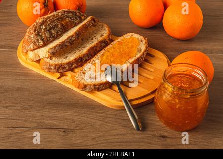 Breakfast prepared with bread, orange marmalade and oranges on a rustic wooden table with a spoon to spread and next to it a pot of marmalade Stock Photo