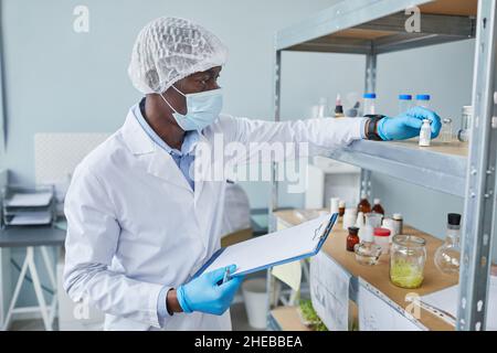 African male doctor in protective mask and white coat working with analysis on the shelves and making notes in the card Stock Photo