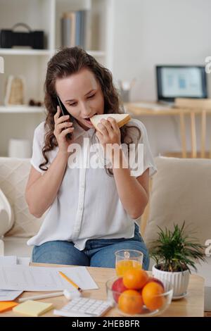 Young female freelance UI designer eating sandwich and talking on phone with client Stock Photo