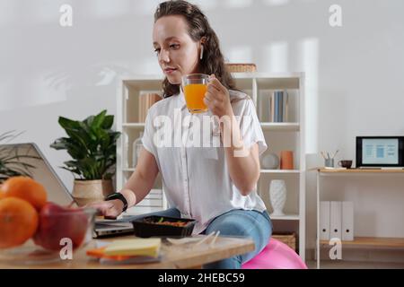 Serious businesswoman drinking cup of juice and checking e-mails from colleagues and clients on laptop when working from home Stock Photo