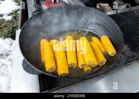 Close-up heap pile of many sweet tasty ripe boiled corn cobs cooked in cast iron cauldron on open fire coal at agricultural food fest fair on winter Stock Photo